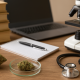 A high-resolution photograph of a cannabis research workspace on a wooden table, featuring a dried cannabis bud, a smaller bud in a glass petri dish, a stethoscope, stacked books, a notebook with a pen, a laptop, and a microscope, all set against a softly lit, neutral background.