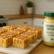 Photorealistic image of cannabis-infused Rice Krispie squares drizzled with caramel and sprinkled with flaky sea salt, arranged on a white cutting board in a clean, stylish home kitchen with warm natural lighting and a jar of infused ghee visible in the background.
