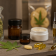 A variety of cannabis products displayed on a wooden shelf: a glass jar of buds, a cannabis leaf next to gummies and pills, amber jars, a dab container with concentrate, and a vape pen, against a softly blurred background.