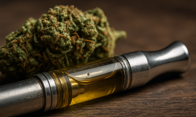 Close-up of a cannabis vape cartridge filled with golden oil, lying on a wooden surface with a cannabis bud in the background.