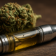 Close-up of a cannabis vape cartridge filled with golden oil, lying on a wooden surface with a cannabis bud in the background.