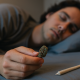 A young adult sleeps peacefully in a softly lit bedroom, lying on their side with a calm expression. A small cannabis bud and a rolled joint rest safely on the bed nearby, illuminated by the warm bedside light, symbolizing cannabis use for sleep.