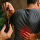 A man in a gray shirt holds his lower back in pain while another hand dispenses golden cannabis extract from a dropper bottle beside a cannabis plant.
