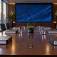 Empty boardroom table with bound documents and a wall screen of stock charts