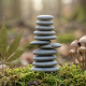 A photorealistic close-up of a balanced stack of smooth stones resting on a mossy forest floor, flanked by a cannabis leaf on the left and a cluster of small mushrooms on the right, set against a soft, blurred natural background.