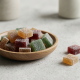 A high-angle, photorealistic shot of assorted square-shaped, sugar-dusted fruit gummies in various colors like amber, deep red, and forest green. Most are gathered in a small, minimalist wooden bowl on a light grey stone surface, with a few gummies and a small green sprig resting nearby in a bright, clean setting.