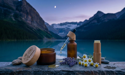 A close-up, photorealistic widescreen photograph of the Canadian CBD skincare collection. On a weathered grey slate slab, three elegant containers are arranged: a frosted amber glass jar of healing salve with a wooden lid, a dark amber serum bottle with a glass dropper, and a textured light-wood balm stick. A single, golden drop of oil is suspended from the dropper, poised above a small pool on the stone. The foreground is accented with fresh sprigs of lavender, several white chamomile flowers, and a few smooth river stones, with a subtle, text-free maple leaf etched into the corner of the slate. The background is a soft-focus vista of a tranquil Canadian glacial lake and snow-capped mountains under a deep blue twilight sky with a sliver of a crescent moon.