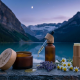 A close-up, photorealistic widescreen photograph of the Canadian CBD skincare collection. On a weathered grey slate slab, three elegant containers are arranged: a frosted amber glass jar of healing salve with a wooden lid, a dark amber serum bottle with a glass dropper, and a textured light-wood balm stick. A single, golden drop of oil is suspended from the dropper, poised above a small pool on the stone. The foreground is accented with fresh sprigs of lavender, several white chamomile flowers, and a few smooth river stones, with a subtle, text-free maple leaf etched into the corner of the slate. The background is a soft-focus vista of a tranquil Canadian glacial lake and snow-capped mountains under a deep blue twilight sky with a sliver of a crescent moon.