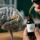 A photorealistic medical illustration featuring a human brain model and a hand holding a cannabis oil dropper, showing a glowing green neural receptor graphic.