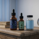 Gemini said A photorealistic, widescreen image of a wooden bedside table featuring three minimalist CBD sleep products: two amber glass dropper bottles with botanical-patterned labels and one clear jar filled with white capsules. The background is soft and airy, showing a blurred window with thin white curtains and a simple round alarm clock, evoking a calm, evening atmosphere.