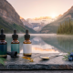 A serene, photorealistic widescreen photograph of three text-free dark amber glass dropper bottles of Canadian CBD oil resting on a grey slate slab and natural wood surface. A glistening sublingual drop of oil is suspended from a glass dropper. The background is a soft-focus vista of a calm, mirror-like lake and a dense forest during the soft, cool blue hour, with the very first warm, golden-pink hues of the sunrise just cresting over distant snow-capped mountain peaks, similar to image_2.png. The air is clear and fresh, with a low mist over the water. The bottles and stone are sharply in focus.