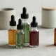 A minimalist, widescreen composition featuring four glass dropper bottles of varying sizes and colors, including amber and clear glass with black caps. The bottles contain liquids in shades of gold, light green, and deep red, arranged neatly on a textured light grey surface alongside neutral-toned ceramic containers and a small green plant.