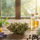 A photorealistic still life on a rustic wooden table featuring a central cannabis flower surrounded by natural sources of terpenes, including lemon peels, lavender sprigs, pine needles, and black peppercorns. In the background, glass vials containing golden oil and a white mortar and pestle sit in warm, natural window light, symbolizing the extraction and study of plant compounds.
